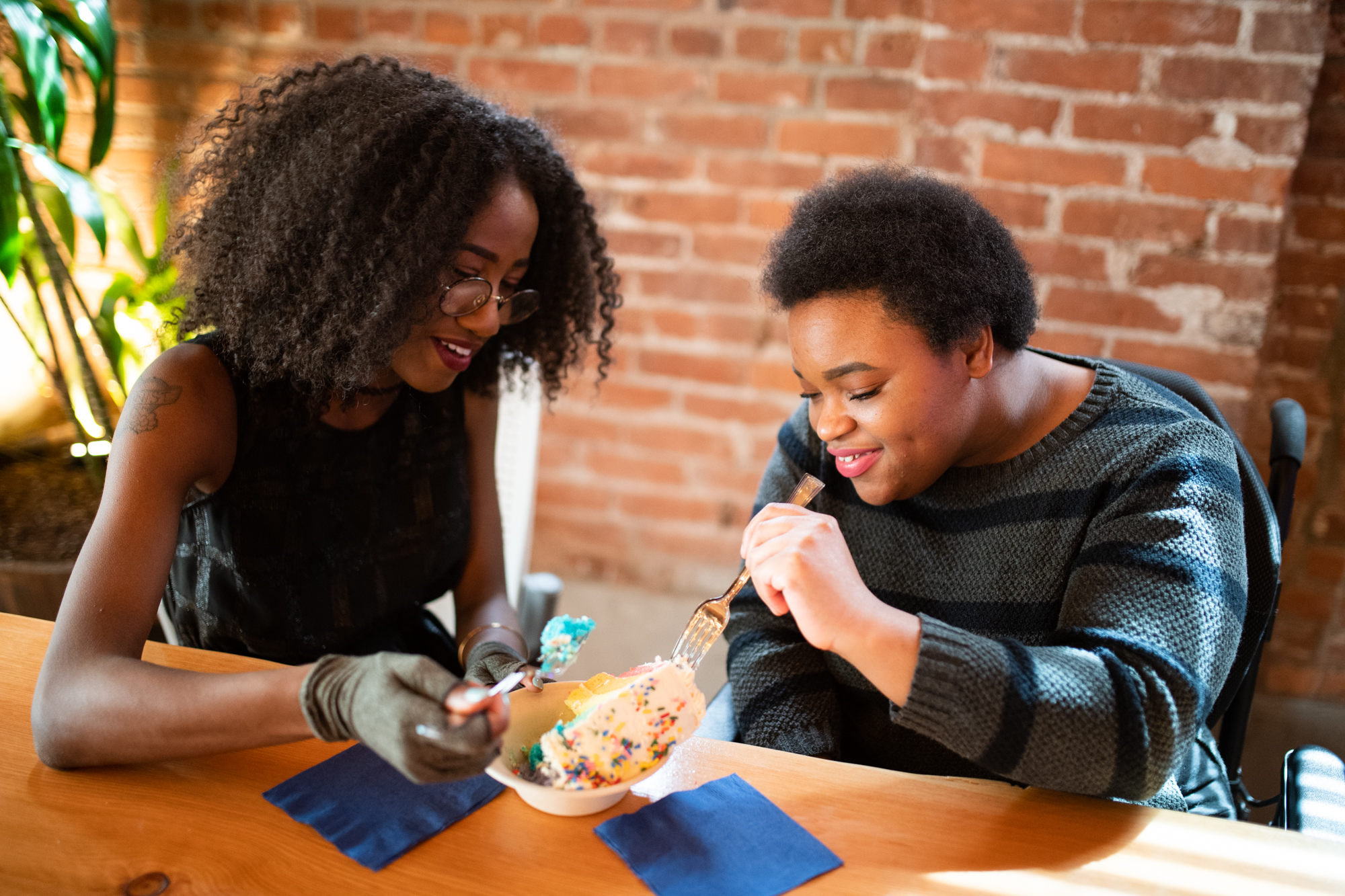 Close-up of two disabled Black people (one woman wearing compression gloves, and the non-binary person in a power wheelchair) sitting next to each other and sharing a slice of cake on a date.