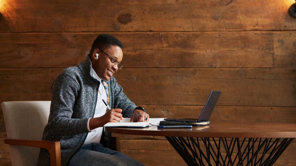 A person wearing a grey sweater and button down sits at a high round table and smiles at their computer monitor as if they were on a video call. 