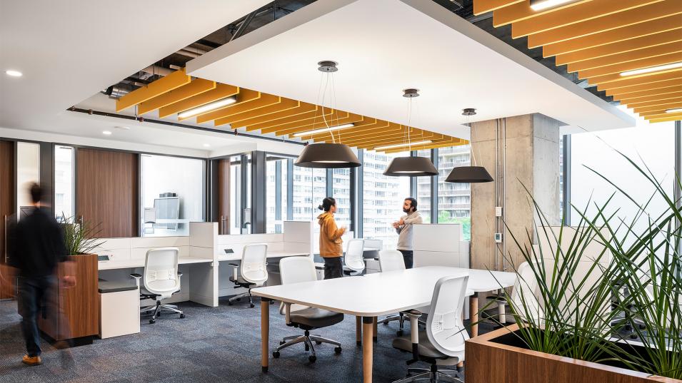 A large room with white tables. On the left of the image, someone is walking away. On the right, two individuals stand talking.